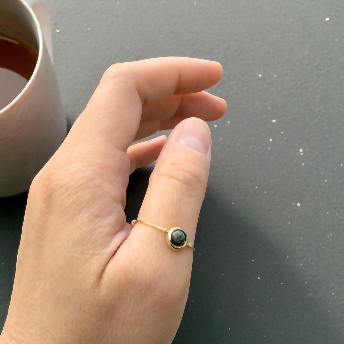 Hand wearing an African Turquoise Gemstone Fidget Ring with a dark stone on a gray surface next to a pink mug.